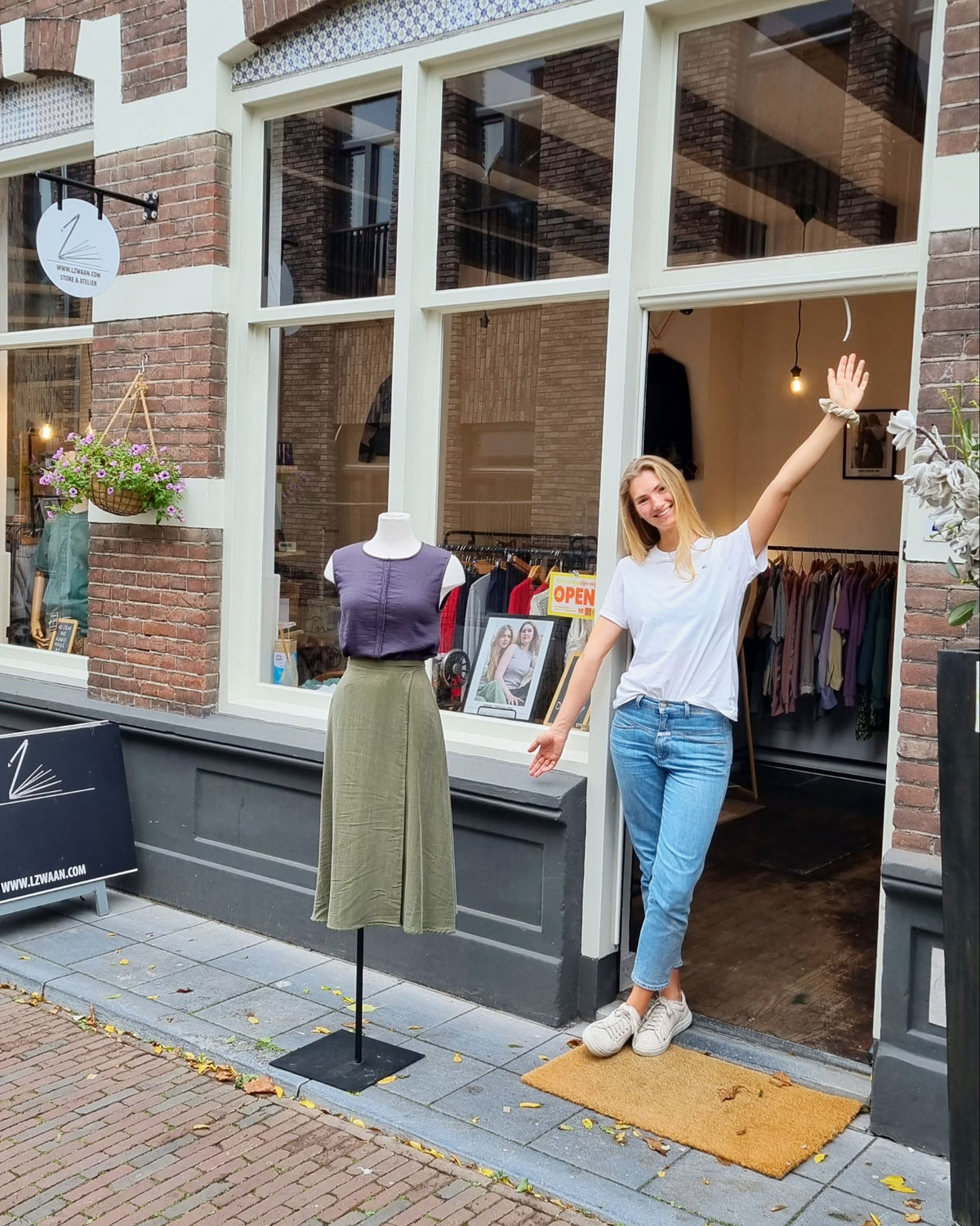 Lara zwaait in de deuropening van de ZWAAN store, naast een paspop in de etalage