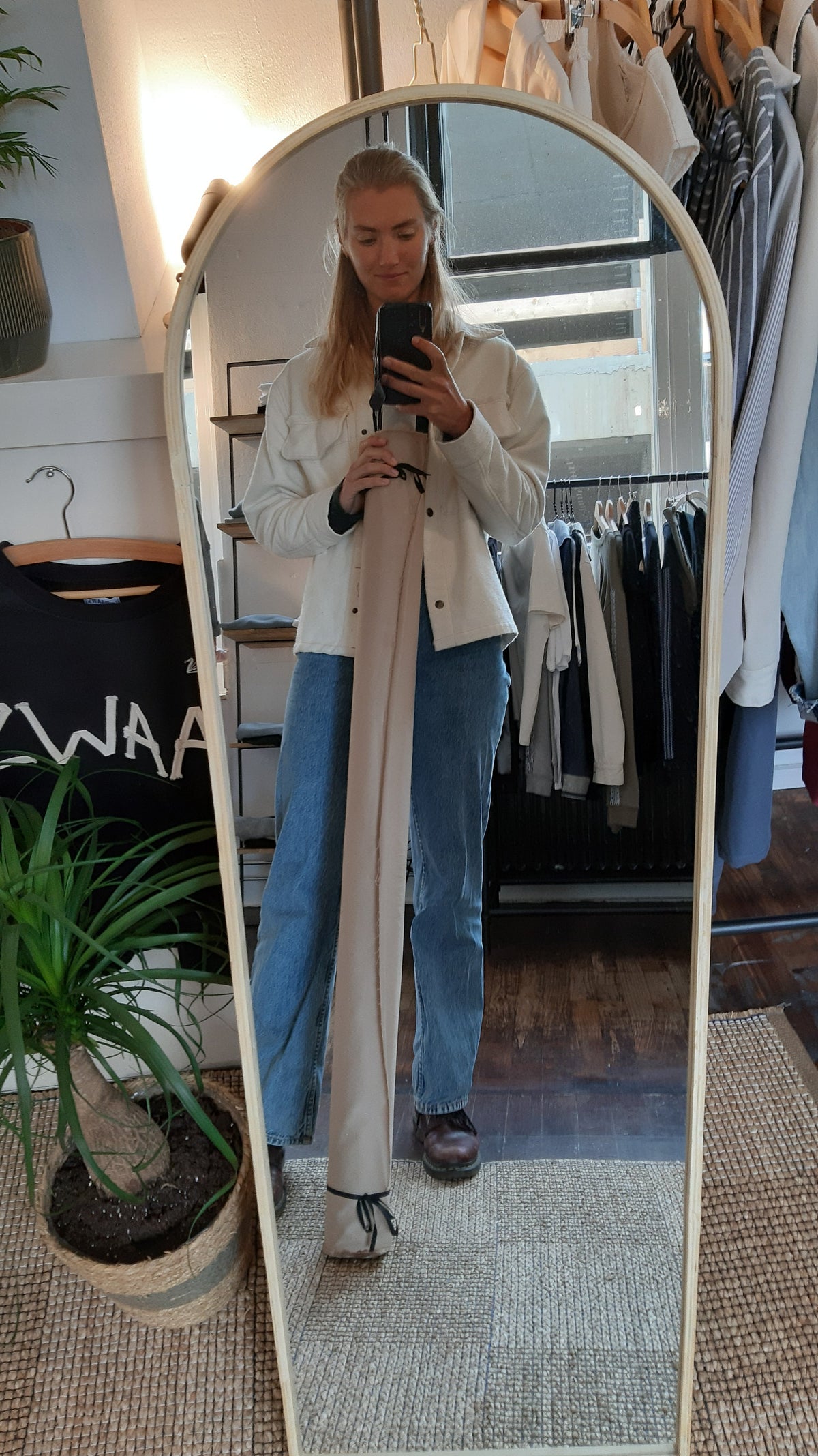 Spiegelselfie van Lara in de ZWAAN store, in een wit jasje en spijkerbroek tussen kledingrekken en planten