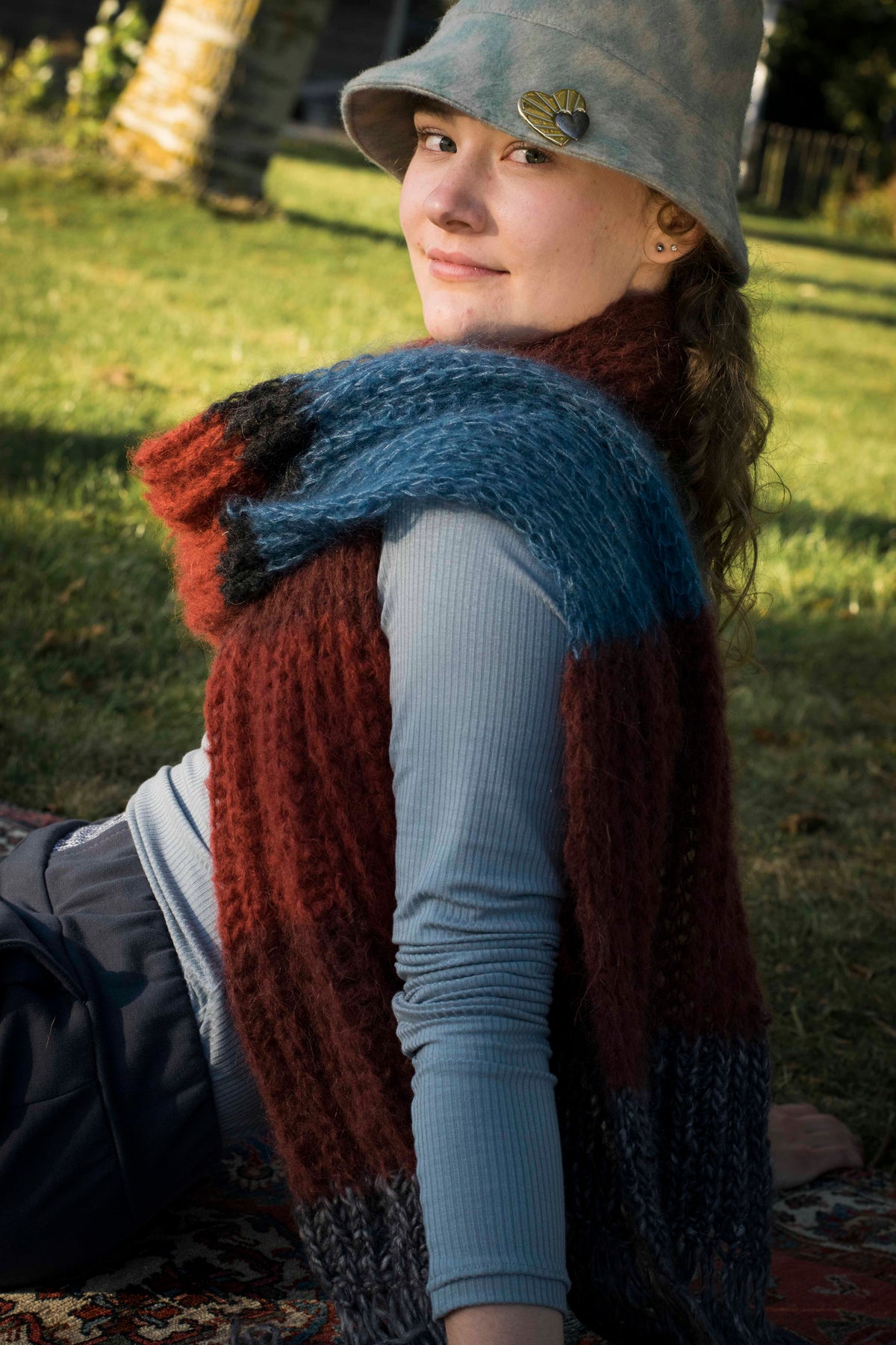 Vrouw in het gras met een handgebreide sjaal in rood, blauw en grijs en een grijze hoed