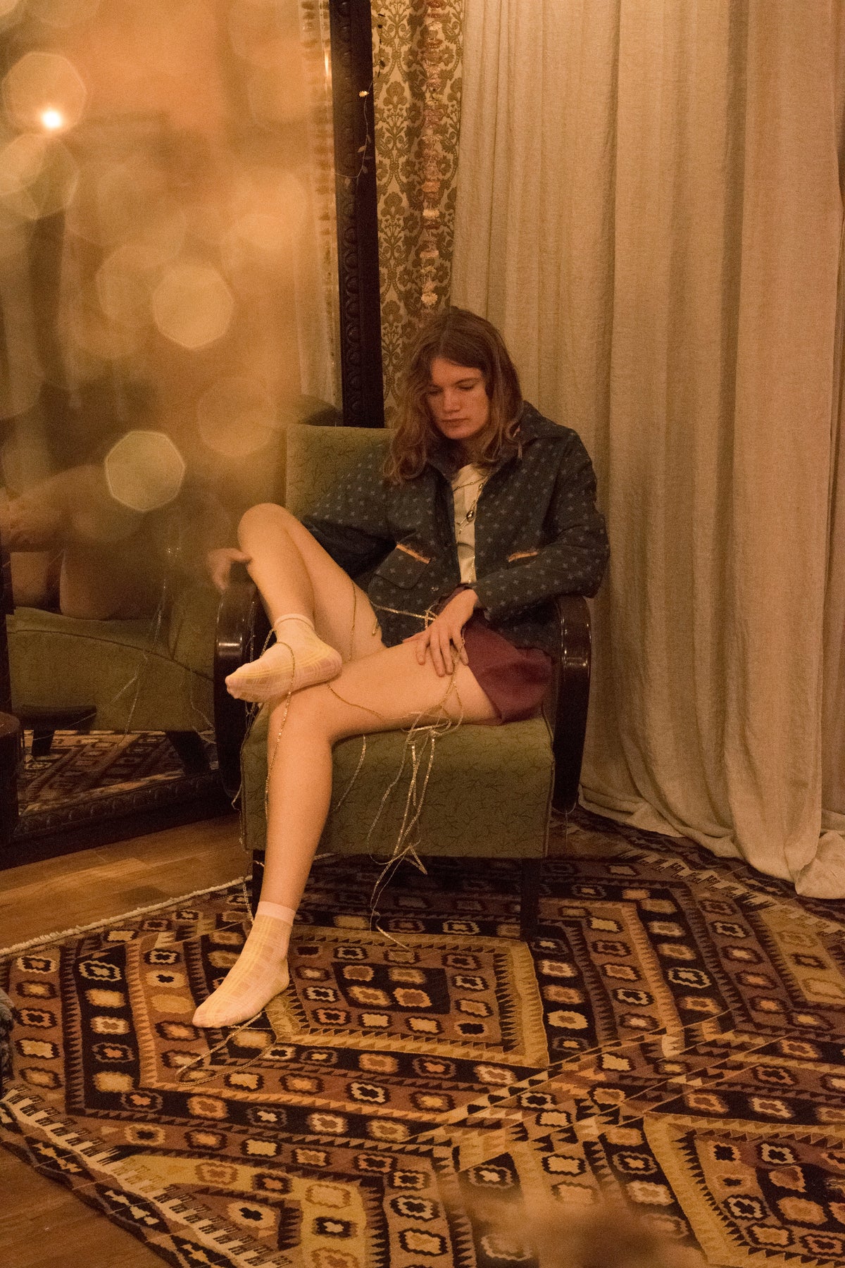 Model met jasje, oudroze short en witte sokken in een groene fauteuil op een oosters tapijt