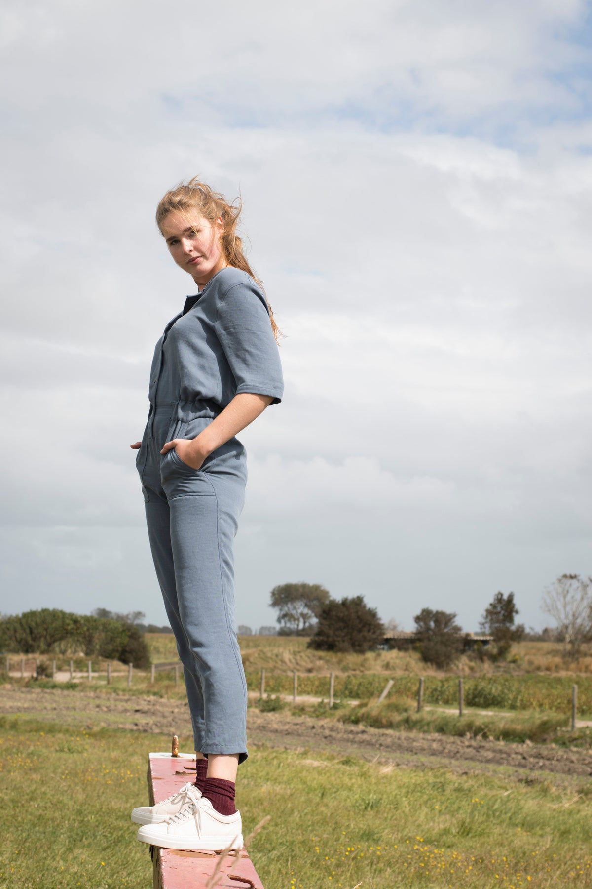 Model in een blauwe jumpsuit en lichte sneakers, staand op een paaltje in een weiland